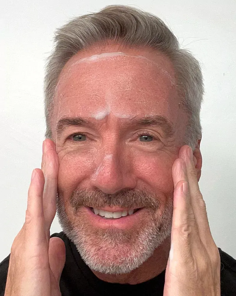 Middle-aged man applying cream to his face, smiling.